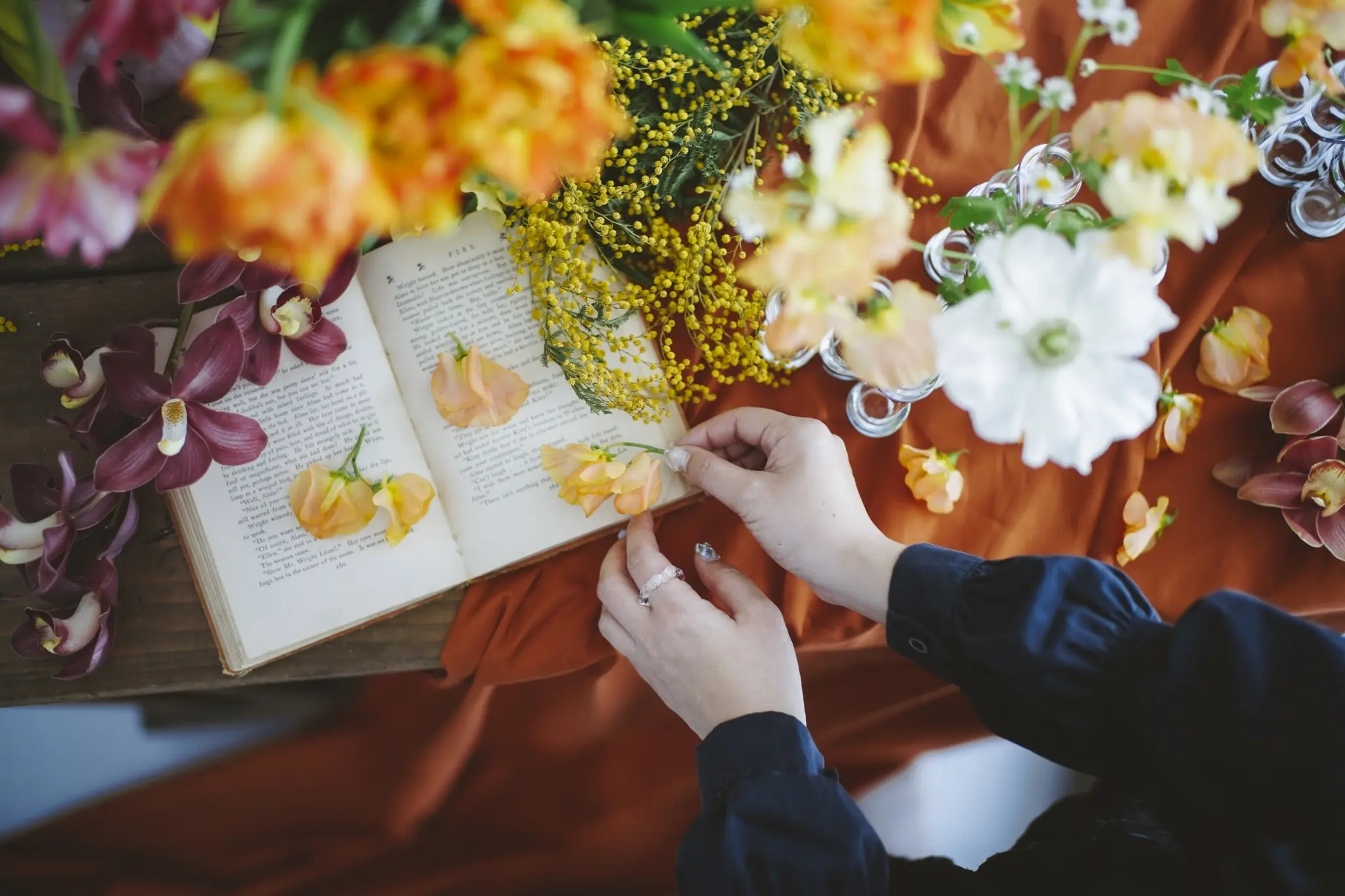 開いた洋書のページにオレンジや黄色の花々とミモザを散りばめ、女性の手がそっとブーケを飾るシーンを捉えたスタイリング写真で、沖縄 フォトウェディングの彩り豊かな演出を表現しています