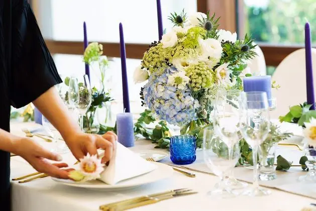 青い紫陽花と白い花を配したセンターピースやキャンドル、グラスが並ぶ披露宴テーブルで、スタッフがゲストのナプキンにデザートをセットしている準備シーン｜テーブルコーディネート