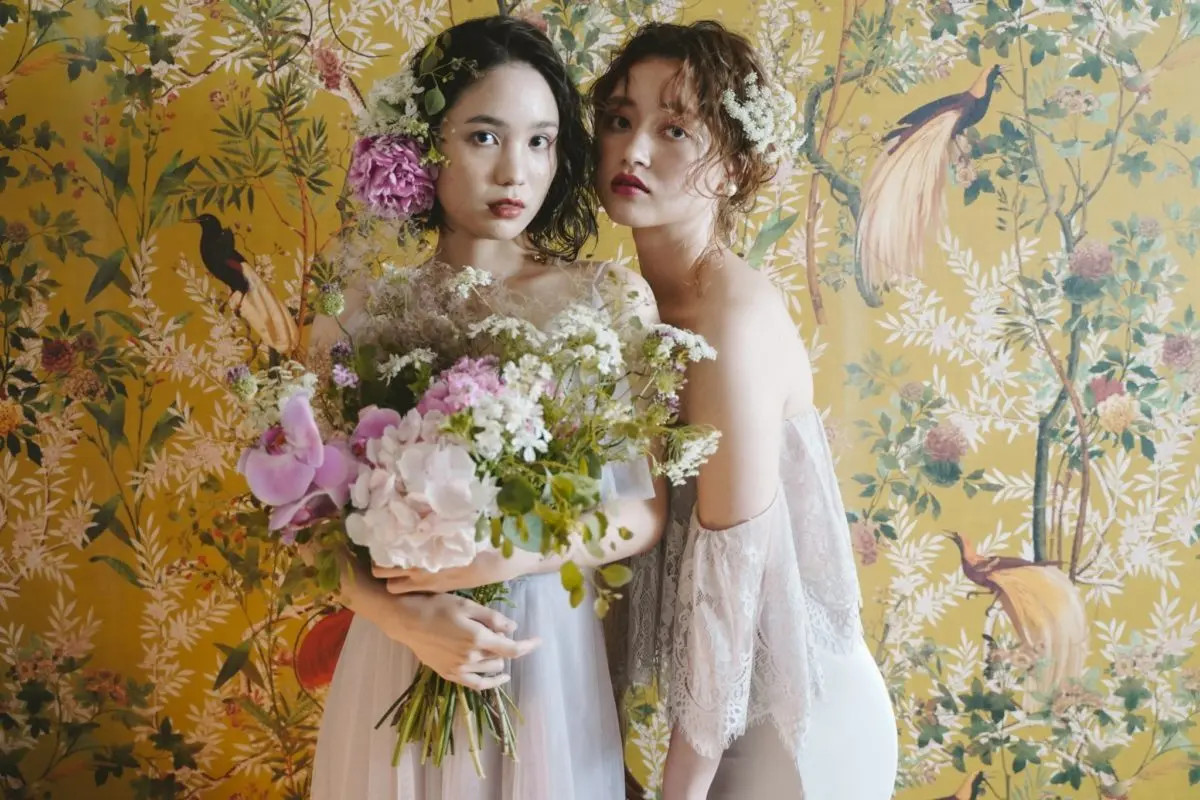 鳥や植物が描かれた華やかな黄色の壁紙の前で、白いドレスの花嫁が大きな花束を抱き、背中の開いたレースドレス姿の女性が寄り添うスタジオ撮影の沖縄 フォトウェディングシーン