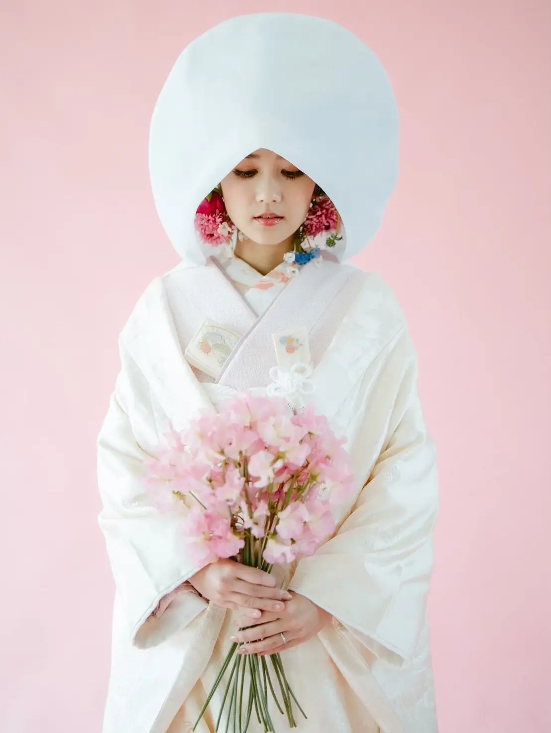 白無垢に綿帽子をまとった花嫁が淡いピンクの背景の前で桜色のブーケを両手に持ち、可憐な耳飾りを添えた凛とした姿が美しい和装写真で、沖縄 フォトウェディングの伝統美とロマンティックな雰囲気を表現しています