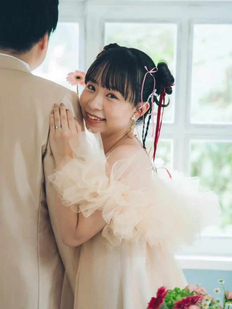 自然な光が入る窓際でベージュのスーツを着た後ろ姿の男性と、レースのドレスを着た女性が並び、女性は手にピンクのガーベラの花を持って笑顔でカメラを見る可愛らしいカップルフォト